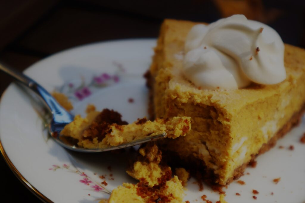 slice of pumpkin cheesecake with a bite next to a fork on a white small china plate lined with purple flowers