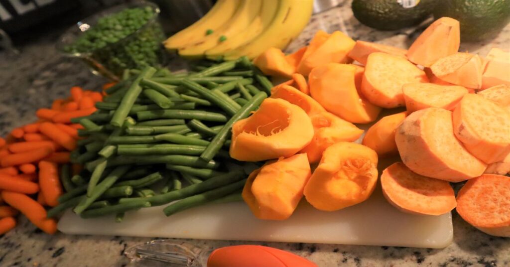 Carrots, green beans. bananas, pumpkin, sweet potatoes, and sweet peas on gray marble kitchen counter with some vegetables on a white cutting board. Also pictured is a vegetable peeler with orange handle and a knife. Silver storage containers in background.