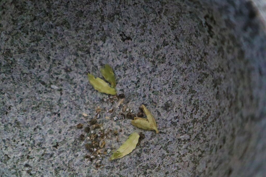 Green cardamom seed husk crushed by mortar in gray and black speckled mortar and black cardamom seeds next to pod husks