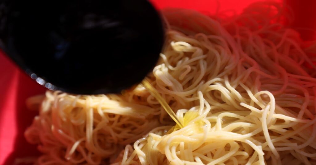 Olive oil poured from black tablespoon onto cooked angel hair pasta that is in a red bowl