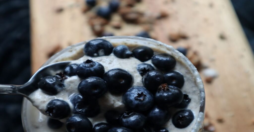 close up of cup with  a lot of bluerries on the spoon. Brown cutting board background with granola and blueberries spread around