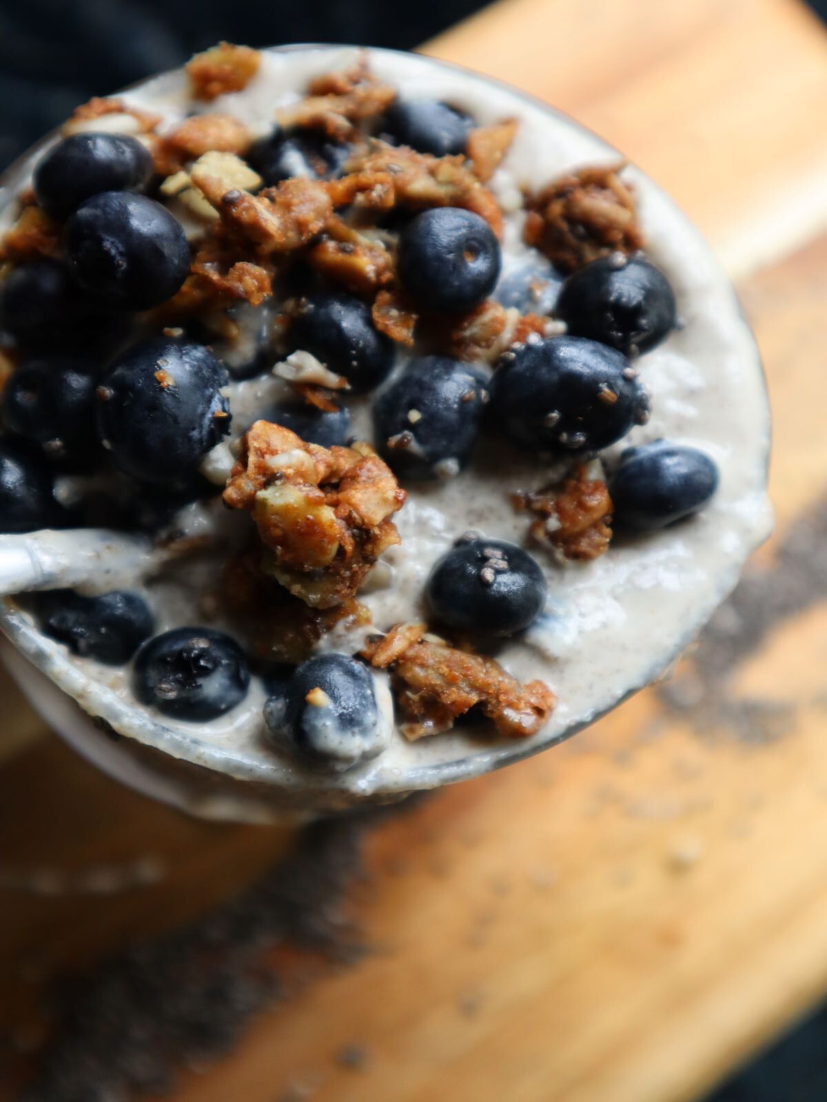 Cup of Chia Seed pudding with spoon in it topped with blueberries and granola on a light brown board background that has chia seeds spread on it