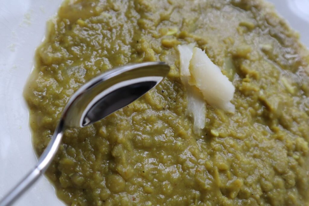 Green Split Pea Soup in a white bowl decorated with slivers of cassava on top the soup. A spoon is near the bowl in the air