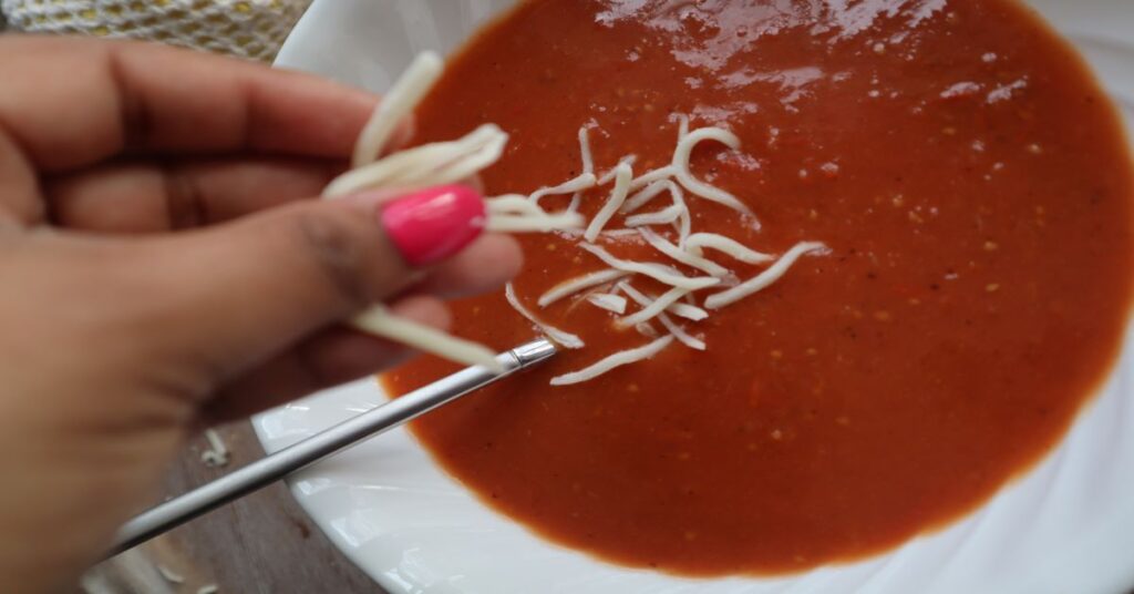 bowl of homemade vegan tomato soup with hand putting shredded mozzarella cheese in it