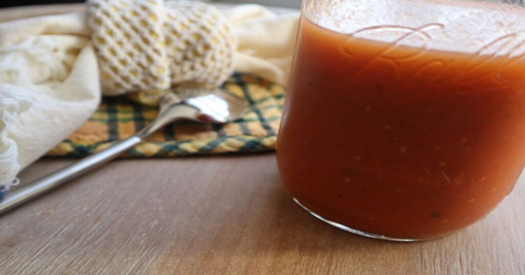homemade tomato soup in mason jar on a cherry wood background with a spoon and green and yellow and yellow and white checkered handcloths in background