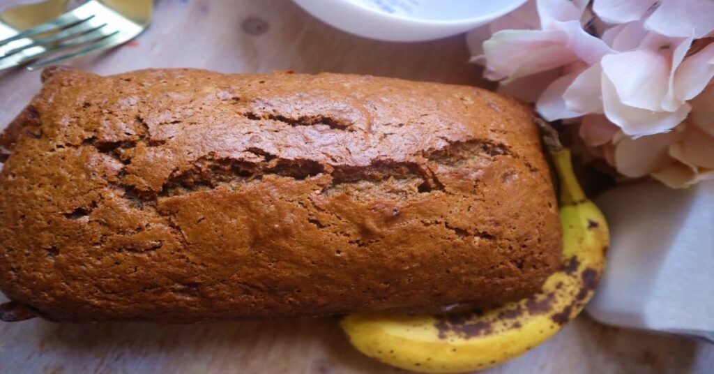 Vegan Banana Bread Loaf uncut displayed with two forks and a banana with flowers as background imagery
