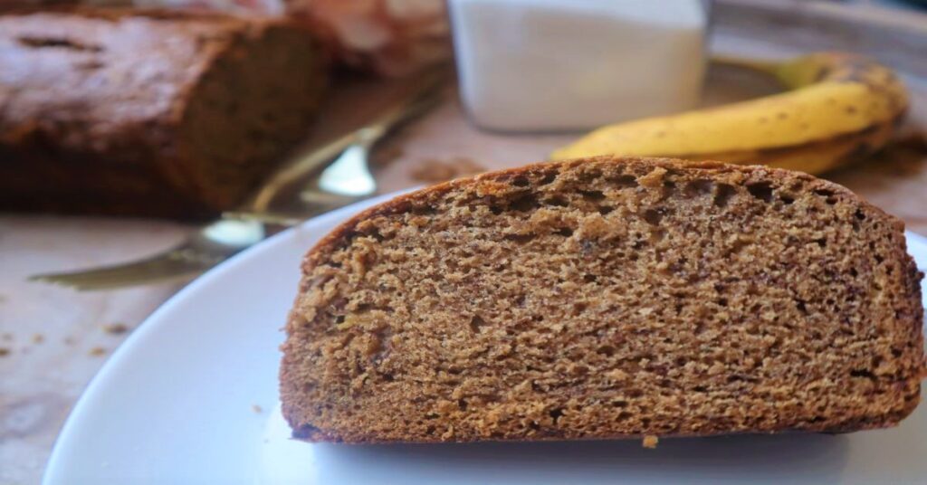 Slice of vegan banana bread on a small white plate. Behind it is a banana, two gold forks, sugar in a clear jar and some pink hydrangeas in a white vase with the rest of loaf