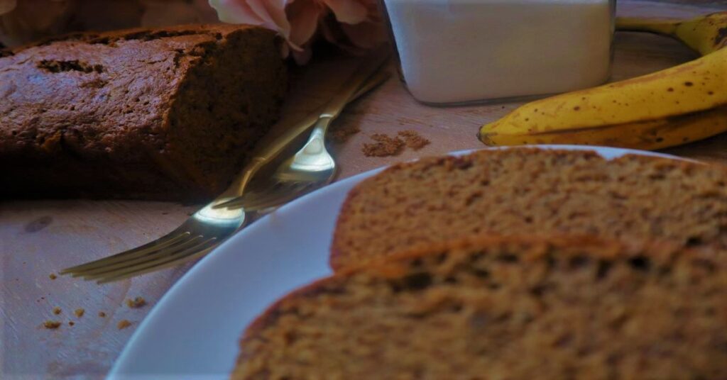 Two slices of vegan banana bread on a small white plate. Behind it is a banana, two gold forks, sugar in a clear jar and some pink hydrangeas in a white vase with the rest of loaf