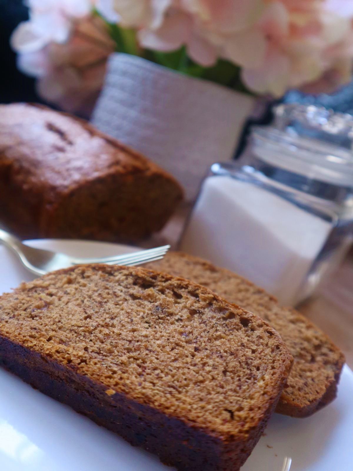 Two slices of vegan banana bread on a small white plate. Behind it is a banana, two gold forks, sugar in a clear jar and some pink hydrangeas in a white vase with the rest of loaf