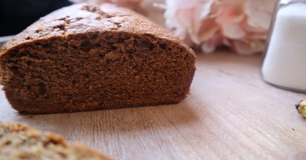 sliced loaf of vegan banana bread on a small white plate. Behind it is a banana, two gold forks, sugar in a clear jar and some pink hydrangeas in a white vase