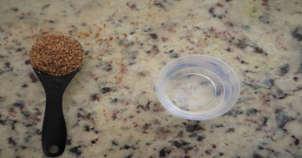 Flaxseed ground in a tablespoon with a ramekin of water next to it on a marble countertop