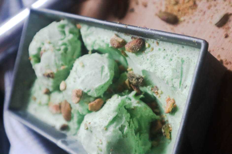 Pistachio Cardamom Ice Cream in gray rectangular container with pistachio pieces on top and ice cream is in three scoops