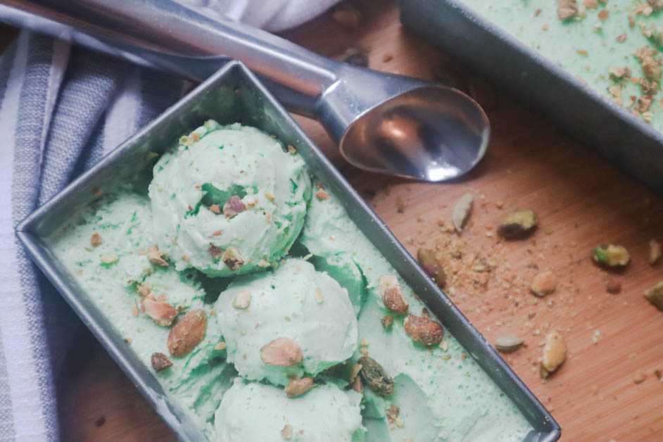 Pistachio Cardamom Ice Cream in gray rectangular container with pistachio pieces on top and ice cream is in three scoops with ice cream scoop displaued on a kitchen towel and part of another ice cream container showing in top right corner