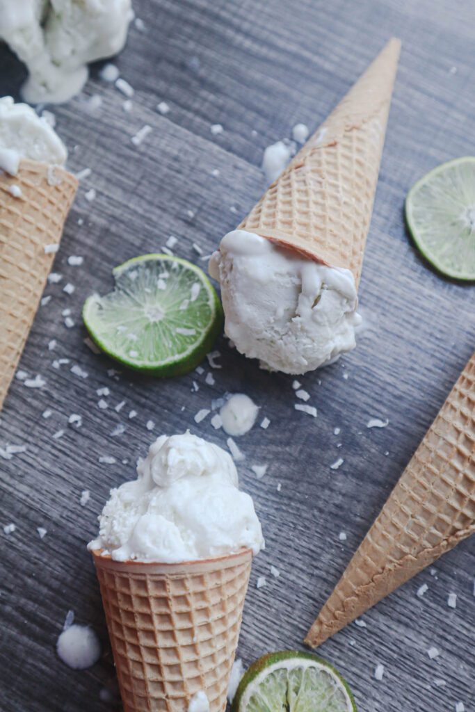 White Scoops of ice cream in waffle cones with sliced limes and shredded coconut on a gray surface