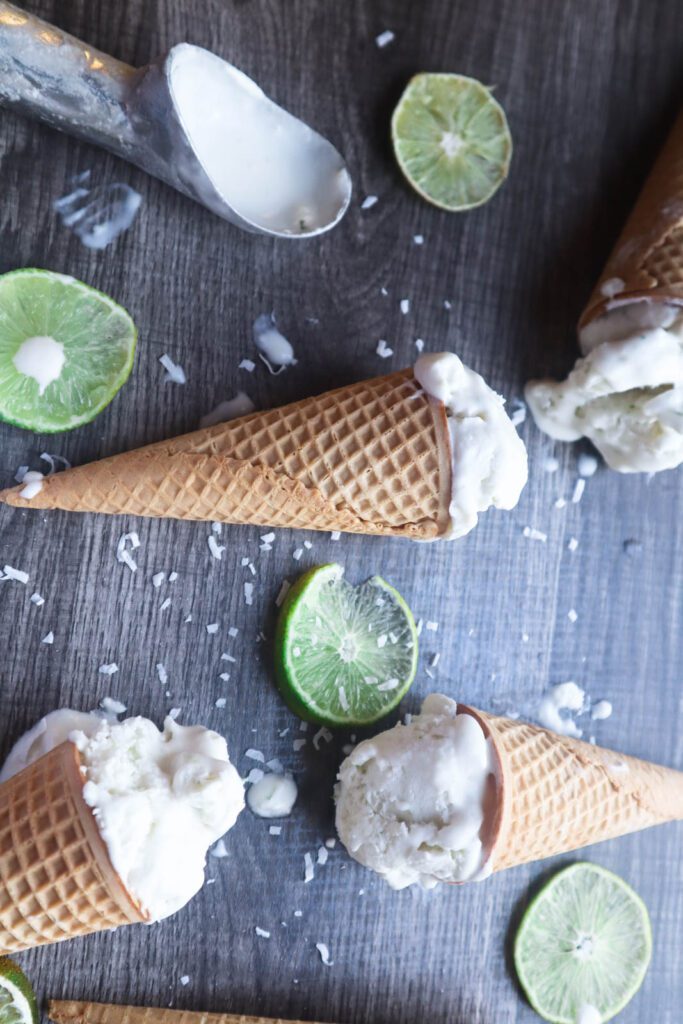 White Scoops of ice cream in waffle cones with sliced limes and shredded coconut on a gray surface and used ice cream scoop