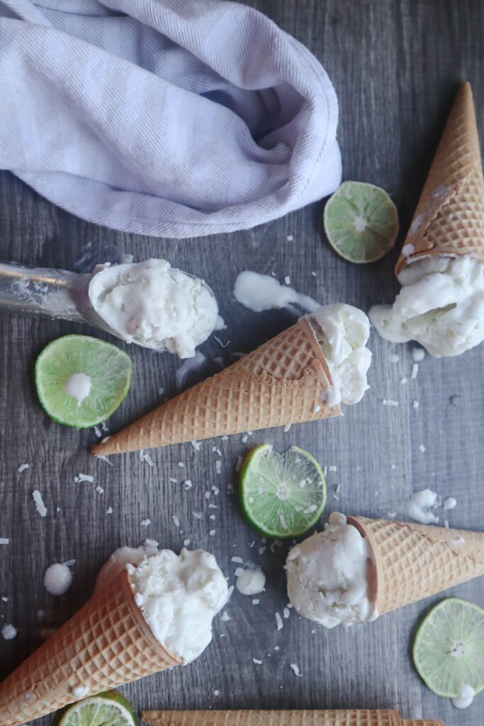 White Scoops of ice cream in waffle cones with sliced limes and shredded coconut on a gray surface. Also scoop of ice cream in ice cream scoop and kitchen towel displayed