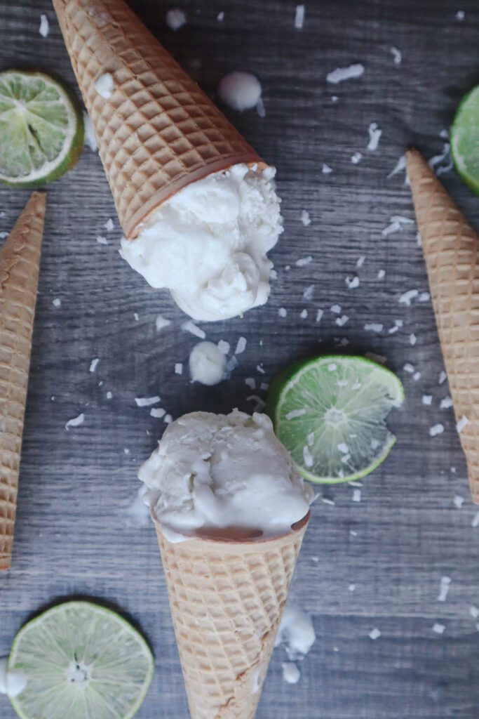 White Scoops of ice cream in waffle cones with sliced limes and shredded coconut on a gray surface