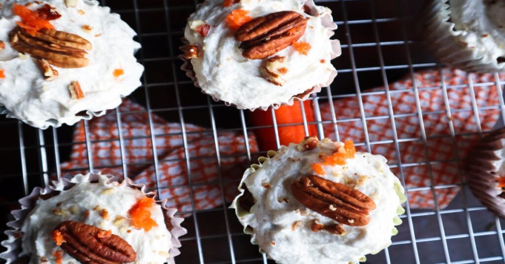 vegan carrot cake muffins with sugar free vegan buttercream frosting on a cooling rack with a carrot and orange and white dish towel below