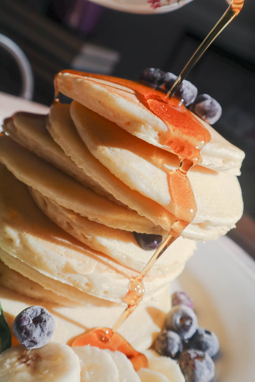 Stack of pancakes with maple syrup being poured onto it. Topped with fresh blueberries