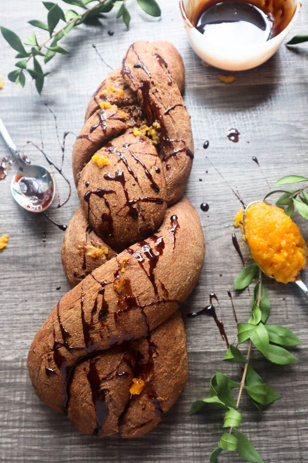 Chocolate Orange Braided Bread drizzled with chocolate syrup on a gray textured background with a silver spoon filled with orange marmalade on it.