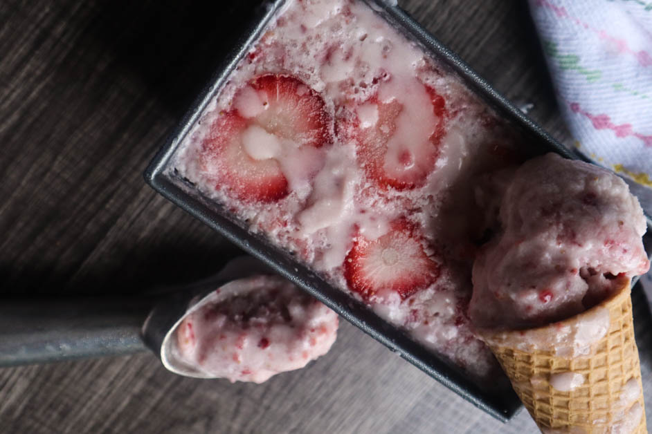 Strawberry ice cream in a waffle cone on top of a container of ice cream with chunks of strawberries pictured in it. A Scoop of ice cream next to container with two waffle cones in the background
