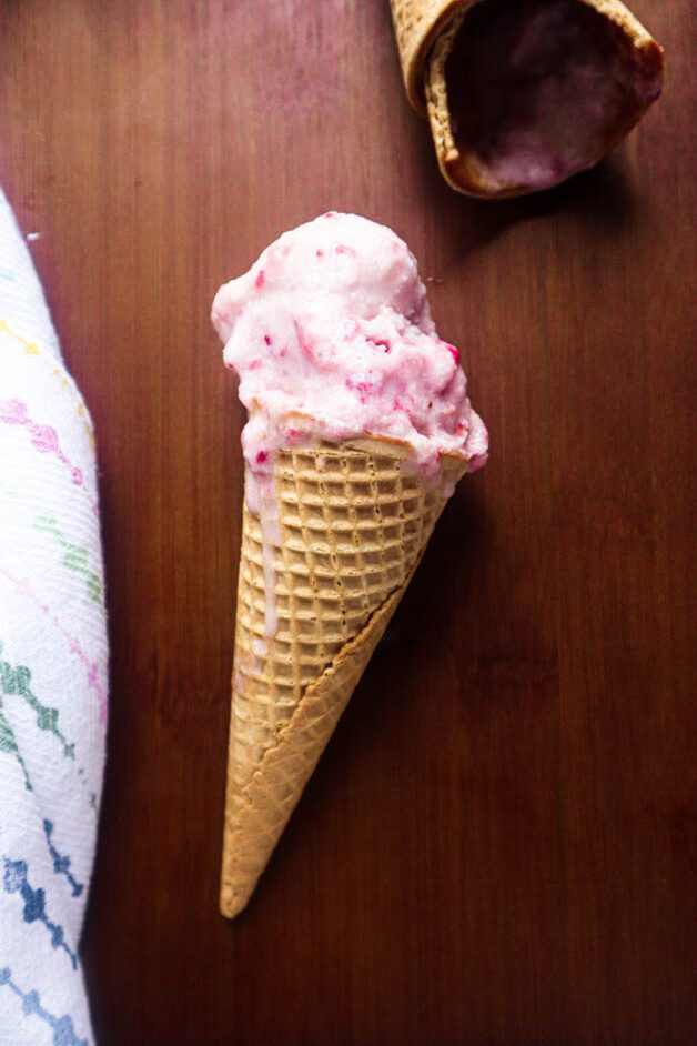 Scoop of strawberry ice cream in a sugar cone with two empty bitten cones near by and a kitchen towel