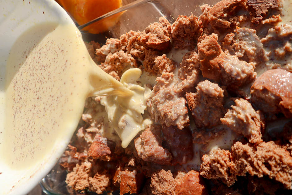 Bread broken in casserole dish with vegan custard in white bowl being poured onto bread and jar of orange marmalade